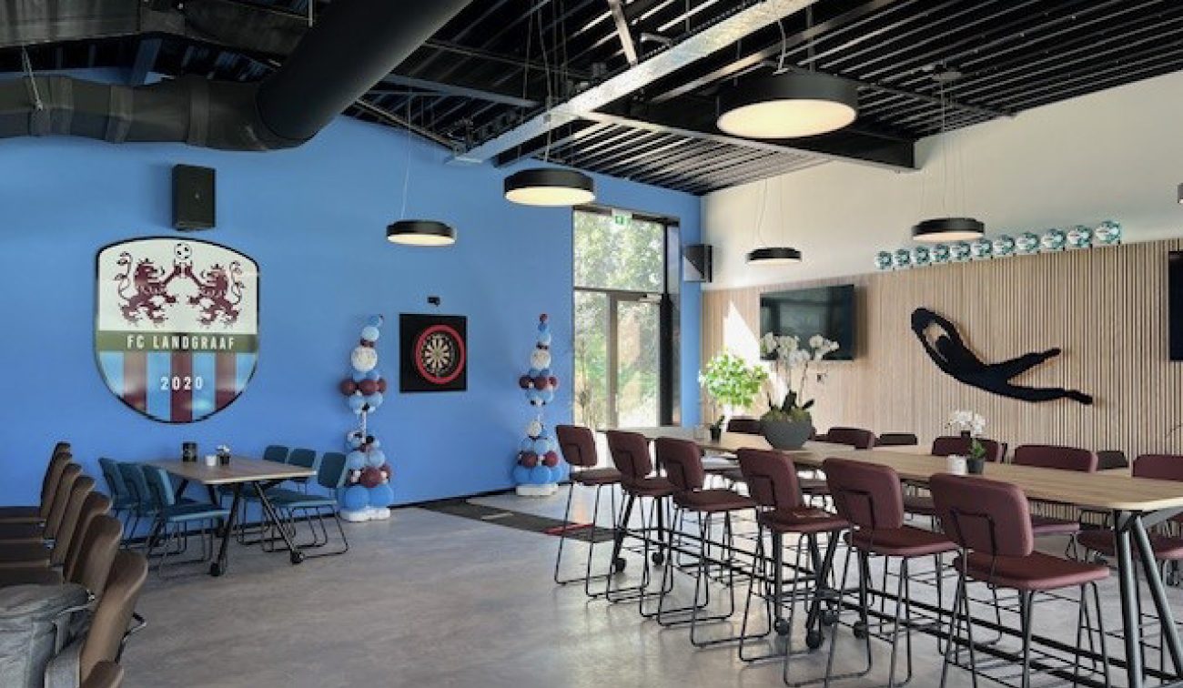 Interieur clubgebouw FC Landgraaf: blauwe wand met clublogo, hoge tafels, barstoelen en sportdecor