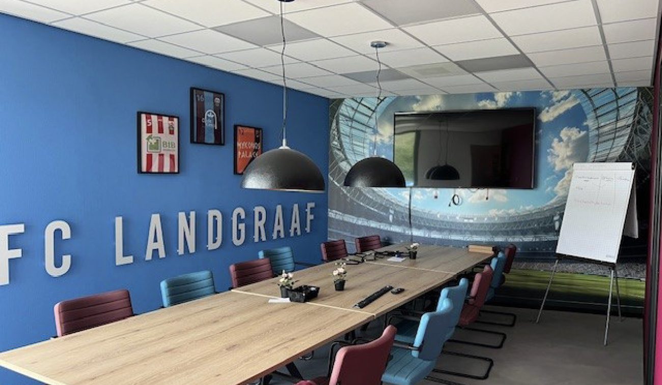 Vergaderruimte FC Landgraaf: lange tafel, design hanglampen, blauwe wand met clubletters en tv-scherm