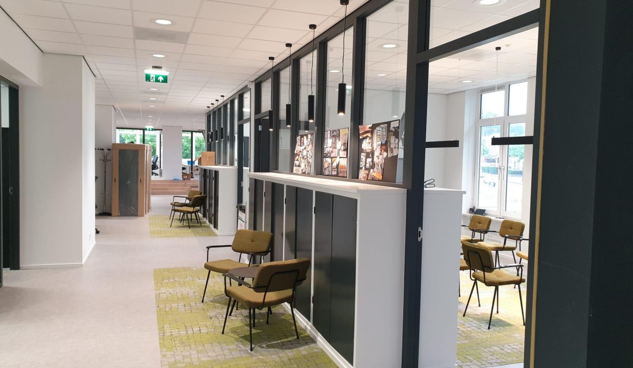 Lichte kantoorruimte met gang en glazen scheidingswanden in zwarte frames; hanglampen langs het plafond, groene fauteuils op tapijttegels en fotopanelen op witte kasten. Grote ramen zorgen voor daglicht.