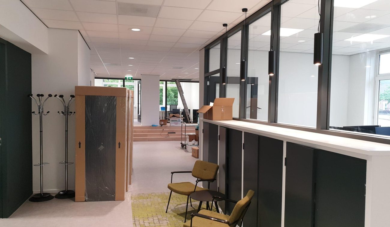 Gerenoveerde kantoorruimte in afbouw: zwarte glazen scheidingswand met hanglampen en hoge kasten, kapstokken en stoelen op een vloerkleed, verhuisdozen; achterin traptreden naar lichte werkruimtes met grote ramen.