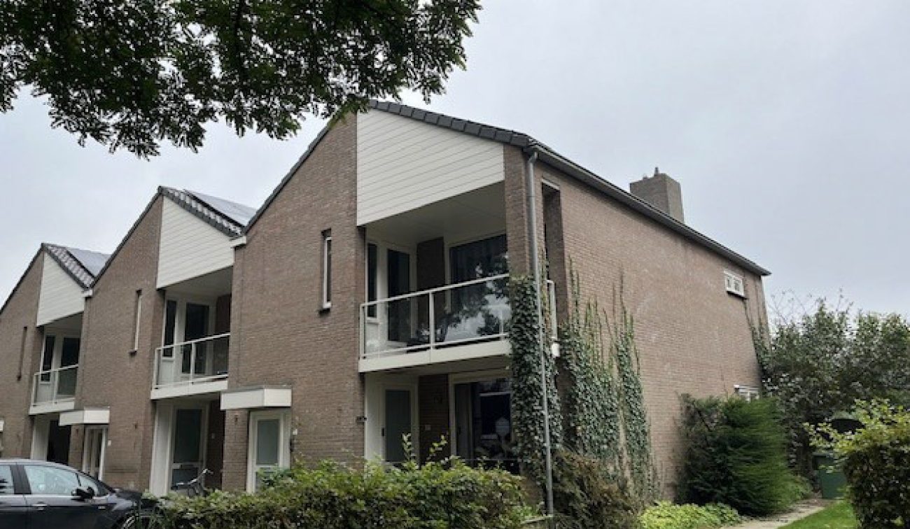 Rij woningen in Stramproy (groot onderhoud) met bakstenen gevels, zadeldaken en witte geveldelen; woningen hebben balkons met glas/white hekwerk, klimplanten langs de gevel en een groen voorterrein onder grijze lucht.