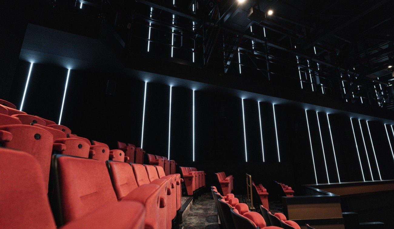 Rij roodpluche theaterstoelen in een donkere zaal met zwarte wanden en verticale LED-lijnverlichting; links de tribune, rechts de rand van het podium/techniekruimte.