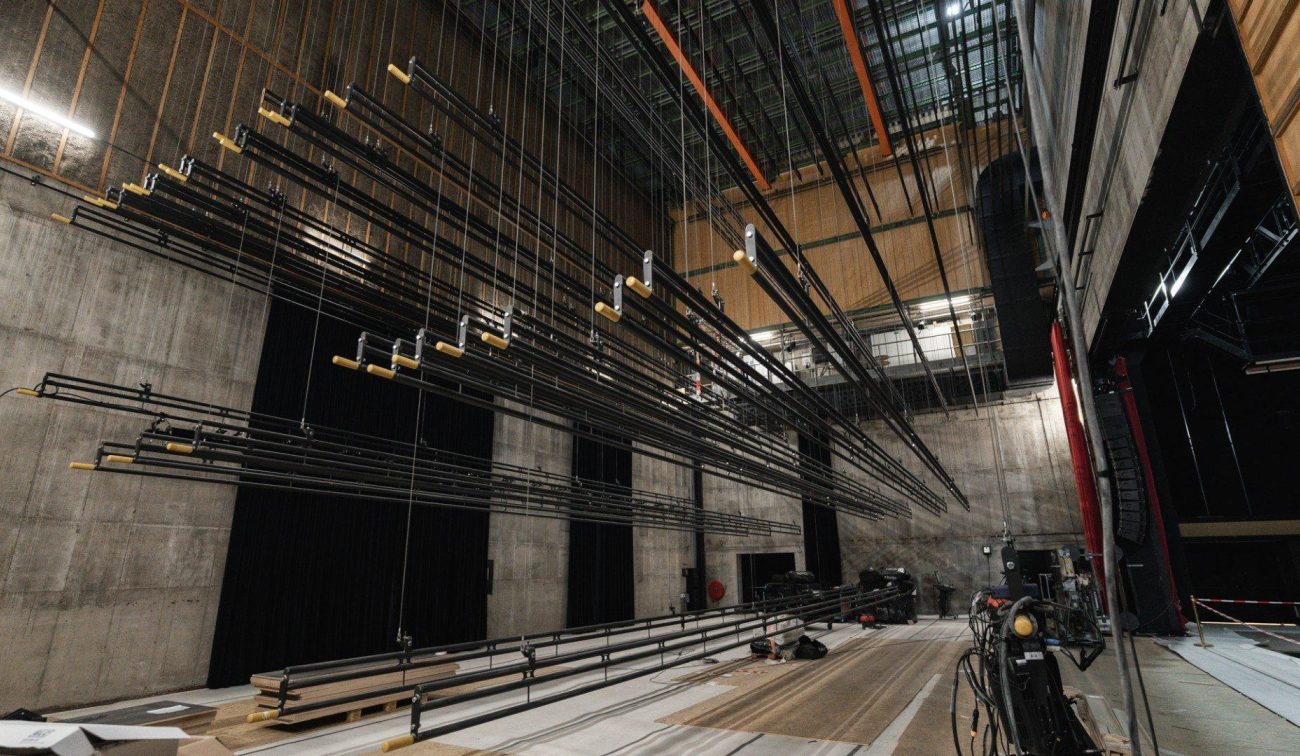 Backstage van Theaterhotel De Oranjerie (Roermond) met hoge toneeltoren: rijen hijsbalken/treklijnen hangen boven een onaf podium, omringd door betonnen wanden, kabels en theatertechnie