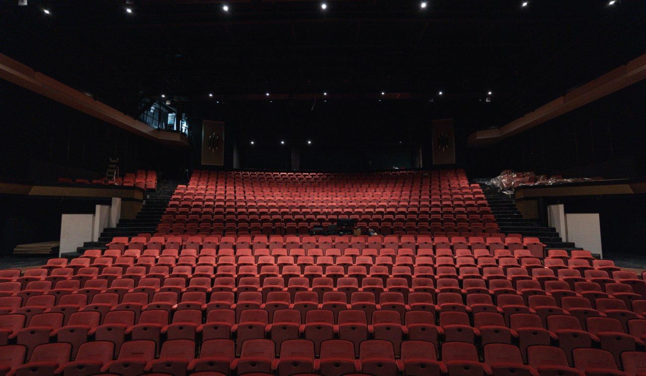 Uitzicht vanaf het podium op een grote theaterzaal met rijen rode stoelen, twee trapgange n aan de zijkanten en een donker plafond met minimale verlichting.