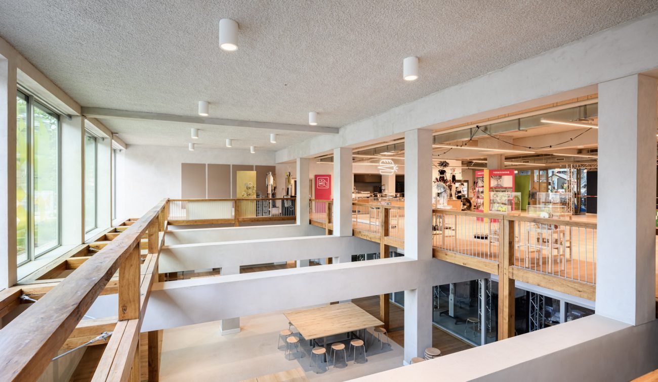 Bovenverdieping rondom vide; witte kolommen, houtaccenten en werkplekken in een open schoolinterieur.