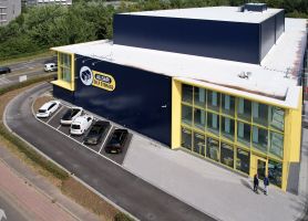 Luchtfoto van de nieuwbouw van Allsafe Self Storage in Maastricht: donkerblauw volume met felgele kaders rond de glazen hoekgevel, witte dakrand en parkeervakken met auto’s langs de voorzijde; twee personen lopen richting de entree, omringd door wegen en groen.