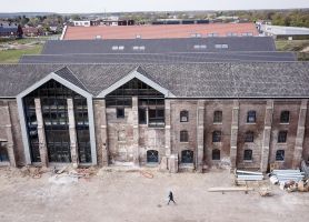 Bakstenen gevel van de Greswarenfabriek in Reuver in renovatie, met grote glazen topgevels; op de voorgrond bouwterrein met materialen en een passerende bouwmedewerker.”