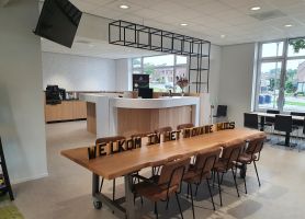 Moderne ontvangstruimte met witte balie en koffiecorner; centraal staat een lange houten tafel met stoelen en de tekst ‘WELKOM IN HET MOVARE HUIS’. Grote ramen en plafondspots zorgen voor veel licht.