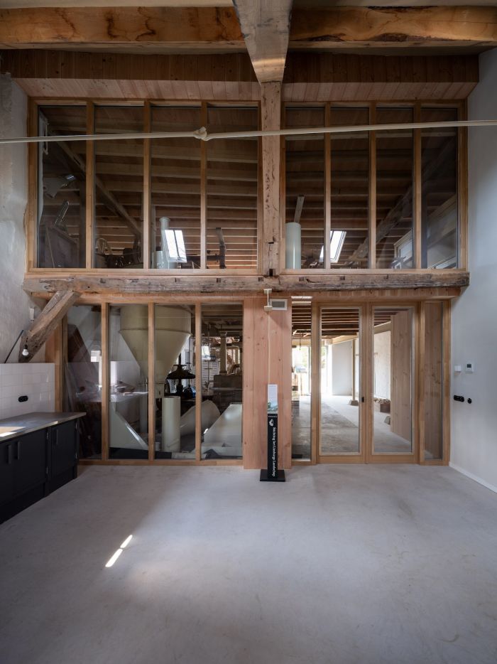 Interieur Grathemermolen met glazen wand waarin de maalinstallatie zichtbaar is (trechters/assen), houten spantconstructie en schuifdeuren naar de gang—combinatie van oud hout en nieuw glas.