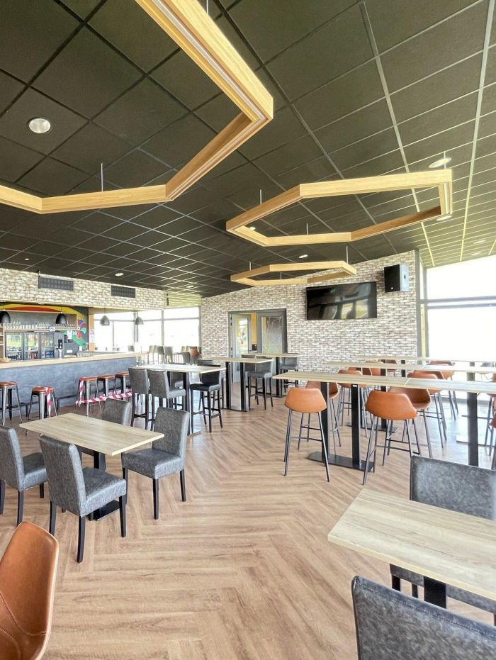 Interieur kantine clubgebouw DVO’20 met bar, hoge en lage tafels, oranje barkrukken, hexagonale houten plafondframes en visgraatvloer; grote tv aan de muur.
