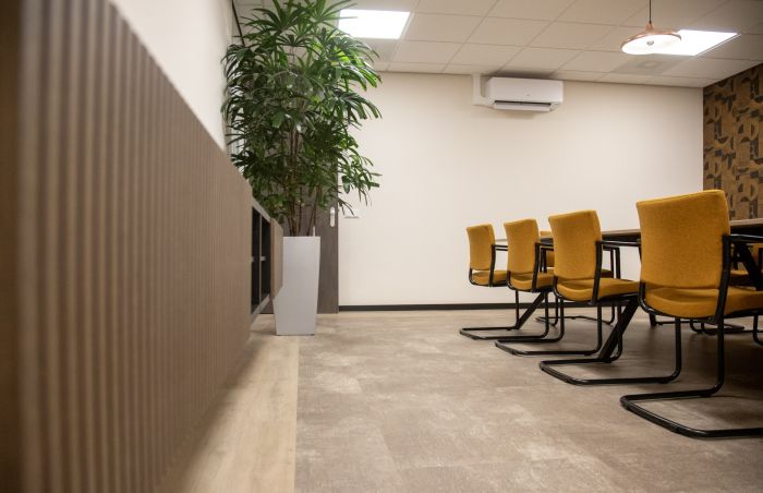 Vergaderruimte met mosterdgele sledestoelen aan een tafel, een hoge kamerplant in een witte pot, lattenwand links, airco-unit hoog aan de muur en neutrale pvc-vloer onder systeemplafond.
