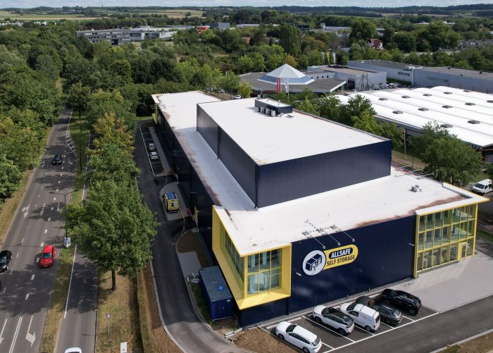 Luchtfoto van de nieuwbouw Allsafe Self Storage in Maastricht: rechthoekig donkerblauw opslaggebouw met wit dak en geel omlijnde glazen hoeken, logo op de gevel, smalle inrit met geparkeerde auto’s en bestelbus, bomen langs de weg en lage bedrijfsgebouwen in de omgeving.