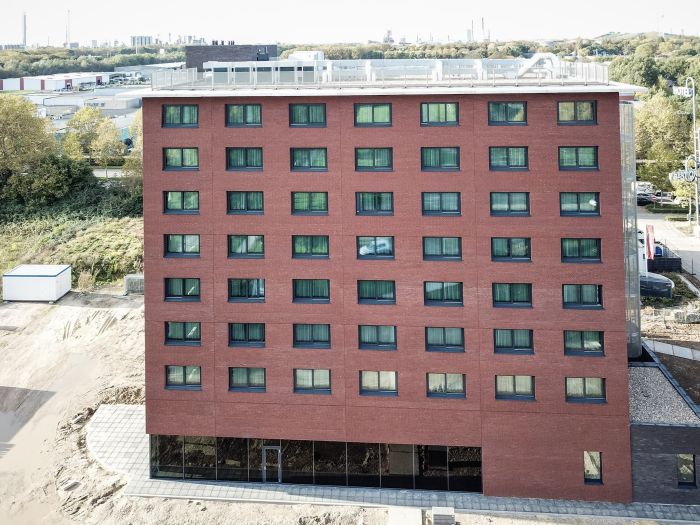 Voorgevel van het nieuwe Bastion Hotel in Geleen: rood­bakstenen blok met een strak raster van donkere, rechthoekige ramen over acht verdiepingen; op het platte dak staan installaties. Aan de voet van het gebouw is nog bouwterrein zichtbaar.