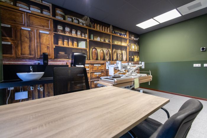 Consultatieruimte met apothekerskast-fotowand, groene accentmuur, wastafel en verstelbare behandeltafel naast bureau