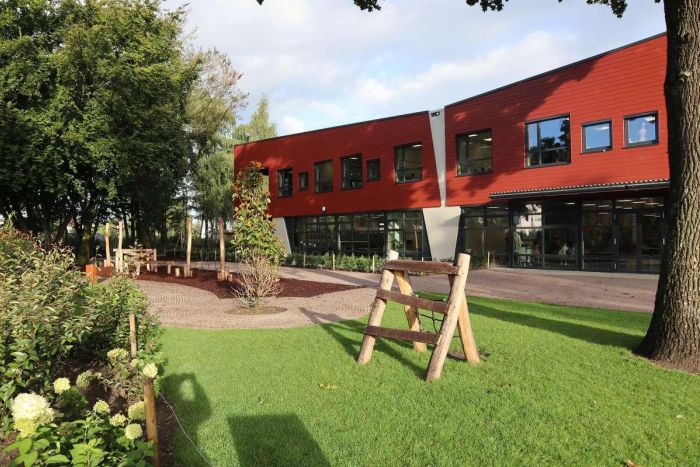 Groen schoolplein met gazon en natuurlijke speeltoestellen van hout; op de achtergrond de rood beklede gevel van basisschool Den Doelhof met grote ramen en glazen entree.