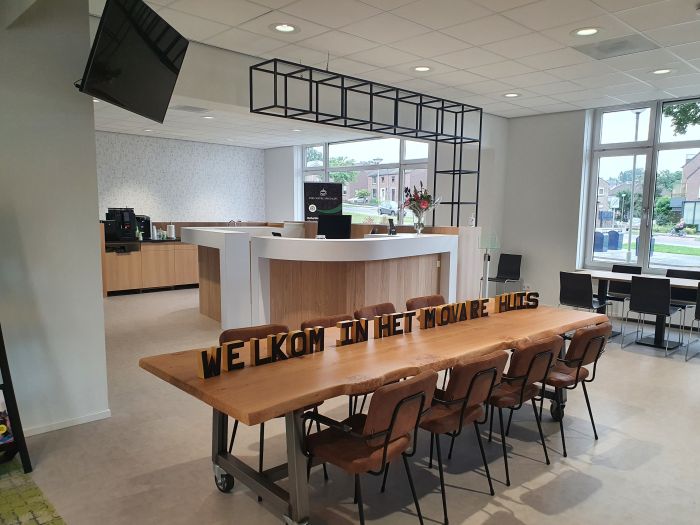 Moderne ontvangstruimte met witte balie en koffiecorner; centraal staat een lange houten tafel met stoelen en de tekst ‘WELKOM IN HET MOVARE HUIS’. Grote ramen en plafondspots zorgen voor veel licht.