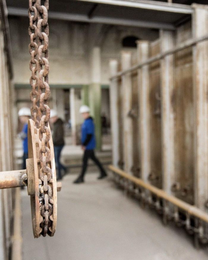 Close-up van een roestige ketting en katrol in een industriële hal; op de achtergrond lopen vervaagde personen met bouwhelmen tussen kolommen.