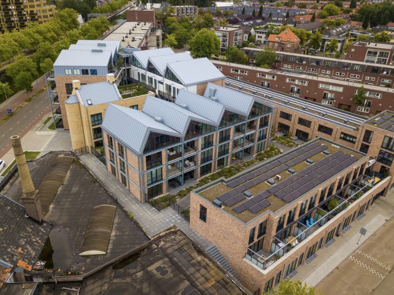 Luchtfoto van modern appartementengebouw met grote ramen, zadeldaken en zonnepanelen op het dak.