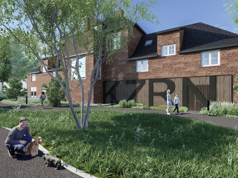 Impressie van de nieuwbouwkazerne in Helmond overdag, met een modern gebouw van rood metselwerk en houten gevel met grote letters ‘KZRN’, omgeven door groen en wandelpaden waar mensen en huisdieren lopen.