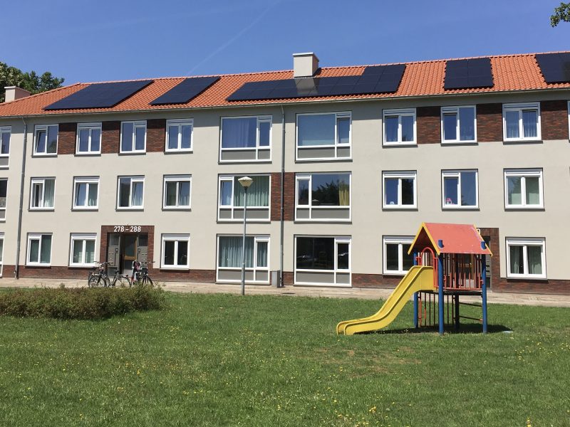 Appartementengebouw met gekleurde gevel en zonnepanelen, met een speeltuin op het grasveld ervoor.