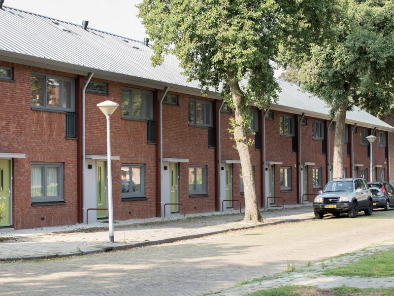 Rij nieuwbouw eengezinswoningen met rode bakstenen gevels en groene voordeuren in een groene woonstraat.