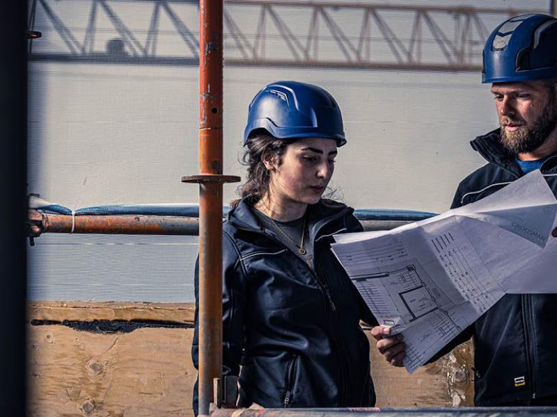 “Twee bouwmedewerkers met blauwe helmen bekijken bouwtekeningen op een bouwplaats.